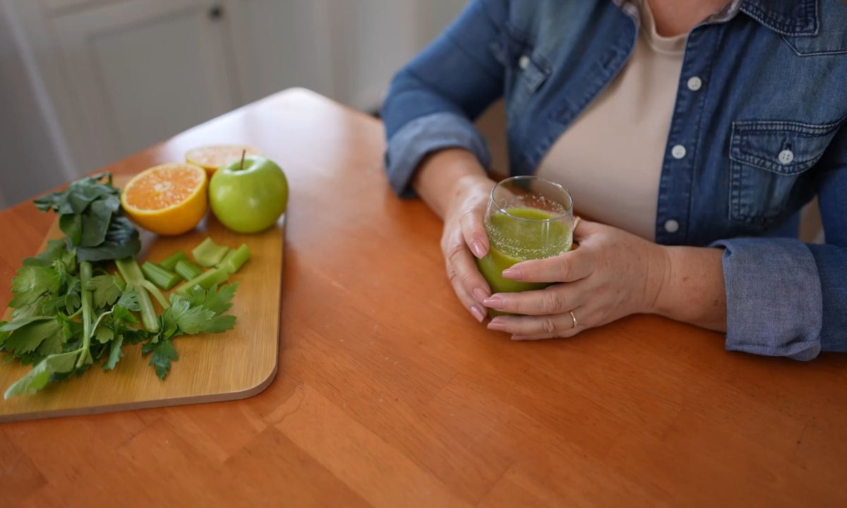 ženska osoba drži čašu sa detoks napitkom, daska na kojoj je pomorandža, zelena jabuka, celer i spanać