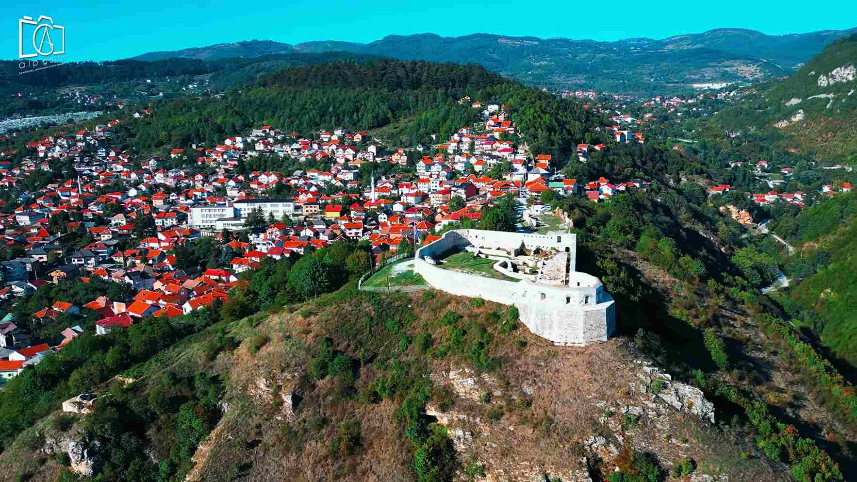 Tvrđava na uzvišenju iznad grada sa crvenim krovovima i planinama u pozadini