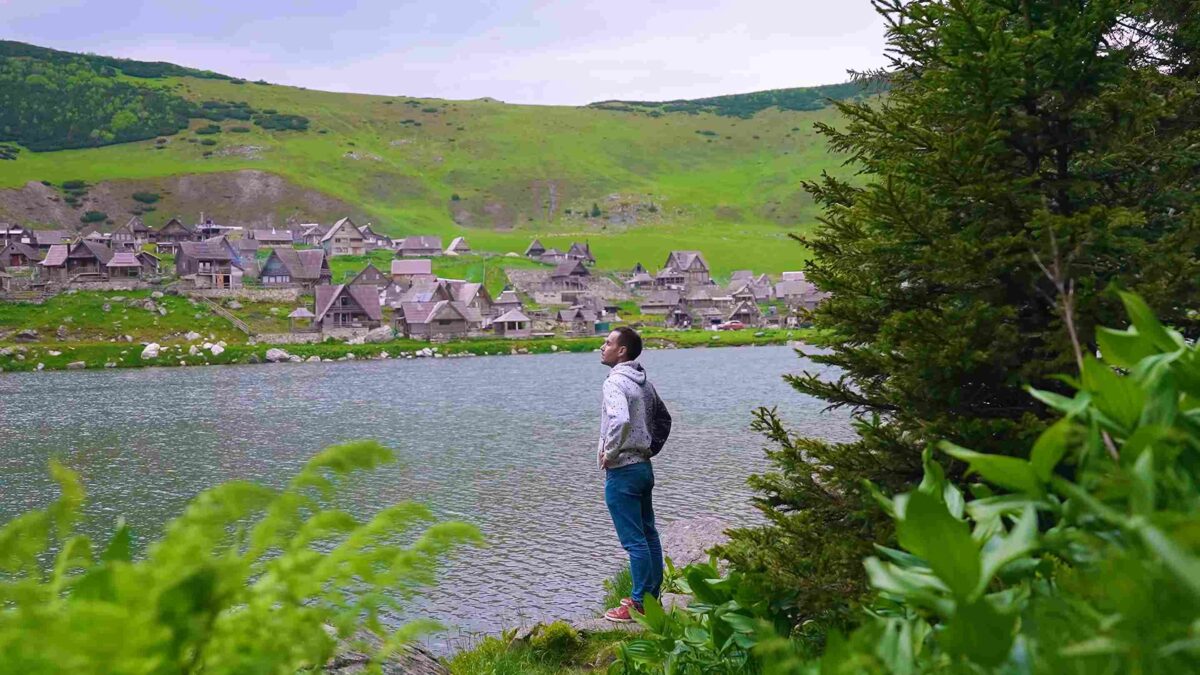Muškarac pored planinskog jezera sa tradicionalnim drvenim kućama i zelenim brdima u pozadini.