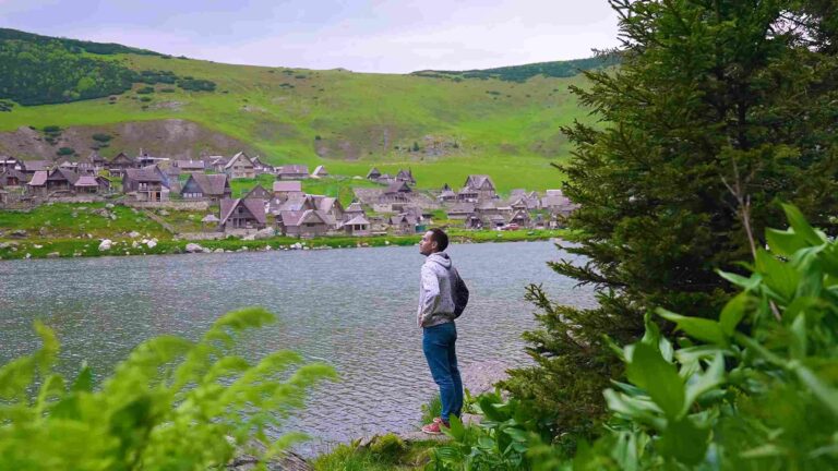 Muškarac pored planinskog jezera sa tradicionalnim drvenim kućama i zelenim brdima u pozadini.