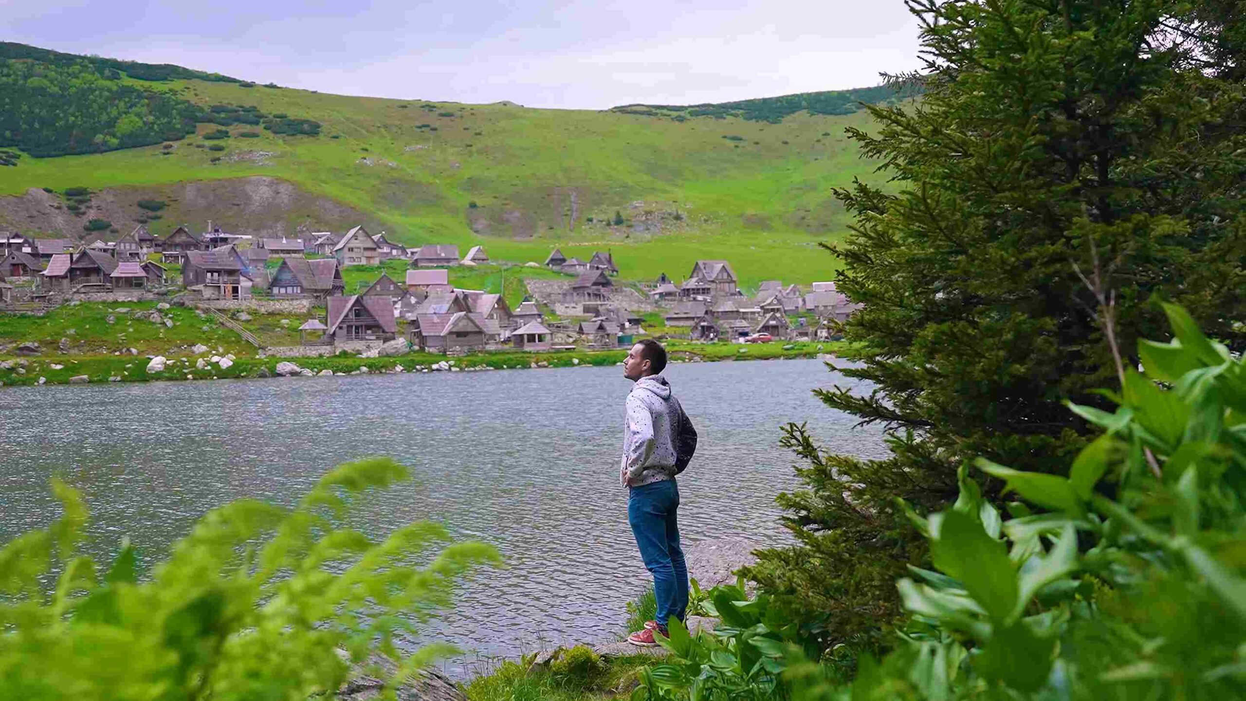 Muškarac pored planinskog jezera sa tradicionalnim drvenim kućama i zelenim brdima u pozadini.